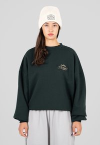 Giovane donna che indossa un berretto lavorato a maglia bianco, una felpa nera oversize con logo e pantaloni grigi chiari, in piedi su uno sfondo neutro.