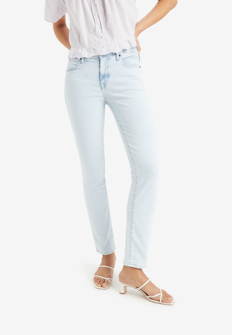 Helle blaue Denim-Jeans mit slim fit und fünf Taschen-Design, kombiniert mit weißen Absatzsandalen. Der Stoff wirkt glatt und leicht dehnbar.