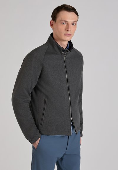 Młody mężczyzna ubrany w ciemnoszarą bluzę zapinaną na zamek błyskawiczny z kieszeniami na zamek oraz niebieskie spodnie, stoi z rękami w kieszeniach na jednolitym tle.