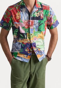 Camicia a maniche corte con bottoni, caratterizzata da un colorato motivo patchwork floreale e tropicale, indossata con pantaloni verde oliva e un orologio da polso.