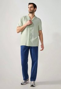 Uomo in piedi che indossa una camicia a maniche corte azzurra con bottoni, jeans blu e sneaker grigie su uno sfondo chiaro e semplice.