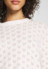 Weißer Strickpullover mit strukturiertem, offenem Muster und blattähnlichen Formen. Gerippter Halsauschnitt mit einem weichen, leichten Gefühl.
