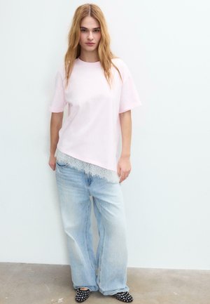 Jeune femme portant un haut rose pâle à manches courtes avec ourlet en dentelle, un jean bleu clair ample et des chaussures noires cloutées, debout contre un mur blanc.