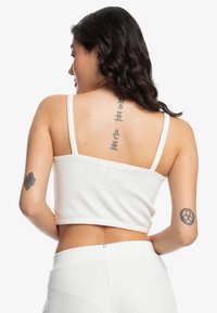 Hvid ribbet crop top med tynde stropper, der har en lige halsudskæring og struktureret pasform. Synlige tatoveringer på ryggen og armen.