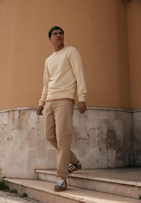 Suéter de punto color beige claro con patrón texturizado, combinado con pantalones beige y zapatillas a rayas marrones, de pie en unos escalones de piedra.