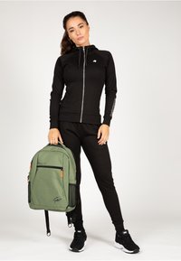 Zwarte zip-up hoodie en joggingbroek, gecombineerd met een groene rugzak met ritsen en mesh zakken. Zwarte sportschoenen maken de look compleet.