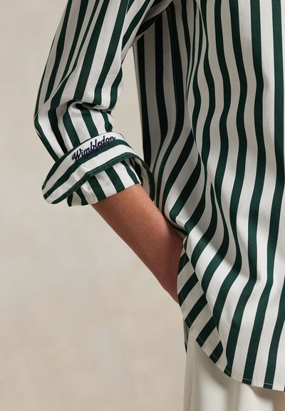 Arm gebeugt mit Hand in der Tasche, trägt ein weißes und dunkelgrün vertikal gestreiftes Hemd mit "Wimbledon" bestickt auf der hochgekrempelten Manschette.