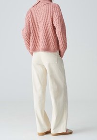 Dzianinowy, oversize'owy różowy sweter o wypukłej teksturze i złożonym kołnierzu, zestawiony z białymi spodniami o szerokich nogawkach i beżowym obuwiem.