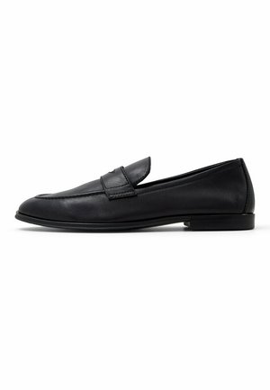 Mocassins en cuir noir avec un bout arrondi, dotés d'une sangle décorative et de détails de couture, reposant sur une semelle basse en caoutchouc noir.