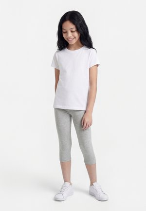 Girl with long black hair wearing a white t-shirt, gray capri leggings, and white sneakers, standing against a white background.