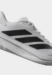 Weiße Freizeitschuhe mit drei schwarzen Streifen. Hergestellt aus Mesh- und synthetischen Materialien, mit einer strukturierten Zwischensohle und einer Gummisohle.
