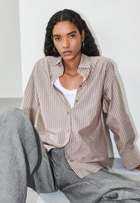 Pruhovaná košeľa na gombíky v svetlohnedej a krémovej farbe, vyrobená z jemnej látky. Kombinovaná s textúrovanými sivými nohavicami s voľným strihom.