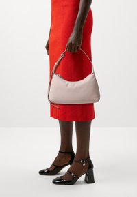 Borsa a tracolla in pelle rosa chiaro con silhouette curva e tracolla grigia, tenuta da una persona che indossa un vestito rosso con texture e tacchi neri lucidi.