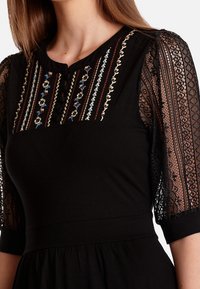 Robe noire avec de courtes manches en dentelle, présentant une broderie multicolore complexe sur le corsage et des détails de boutons au niveau de l'encolure.