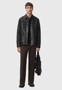 Zwarte leren jas met drukknoopsluitingen en vier voorzakken, gedragen over een zwarte trui, gecombineerd met bruine broek en een zwarte tas.