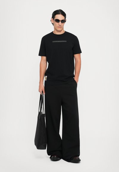 Black cotton t-shirt with short sleeves and tag on hem, paired with wide-leg black trousers. Model holds a black tote bag and wears dark sunglasses.