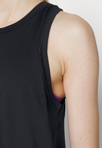 Černý bezrukávový top s kulatým výstřihem a prošívanými okraji, odhalující fialovou podprsenkovou šňůrku pod pravou stranou. Hladká textura.