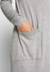 Szary sweter i dzianiny z prążkowanym mankietem i przednią kieszenią o płaskim designie. Tekstura wydaje się miękka i nieco elastyczna.