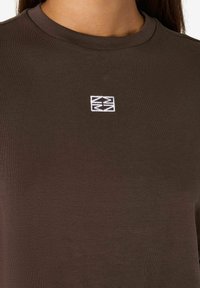 Brūns kokvilnas T-krekls ar apaļu kaklu un baltu izšūtu logotipu uz krūtīm. Gluda tekstūra un brīva piegriezuma.