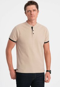 Beige polo tričko s texturovanou tkaninou, černými akcenty na rukávech a knoflíkovou légou. V kombinaci s tmavými džíny.