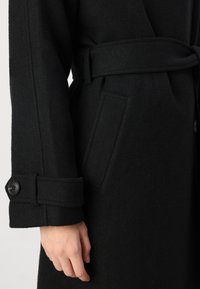 Casaco de lã preto com cinto atado, punhos com botão e um bolso lateral. A textura parece suave e quente, com um design estruturado.