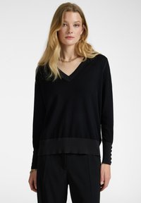 Maglione nero a V con maniche lunghe, scollo e orlo a costine. Presenta dettagli testurizzati ai polsini delle maniche e una vestibilità comoda.