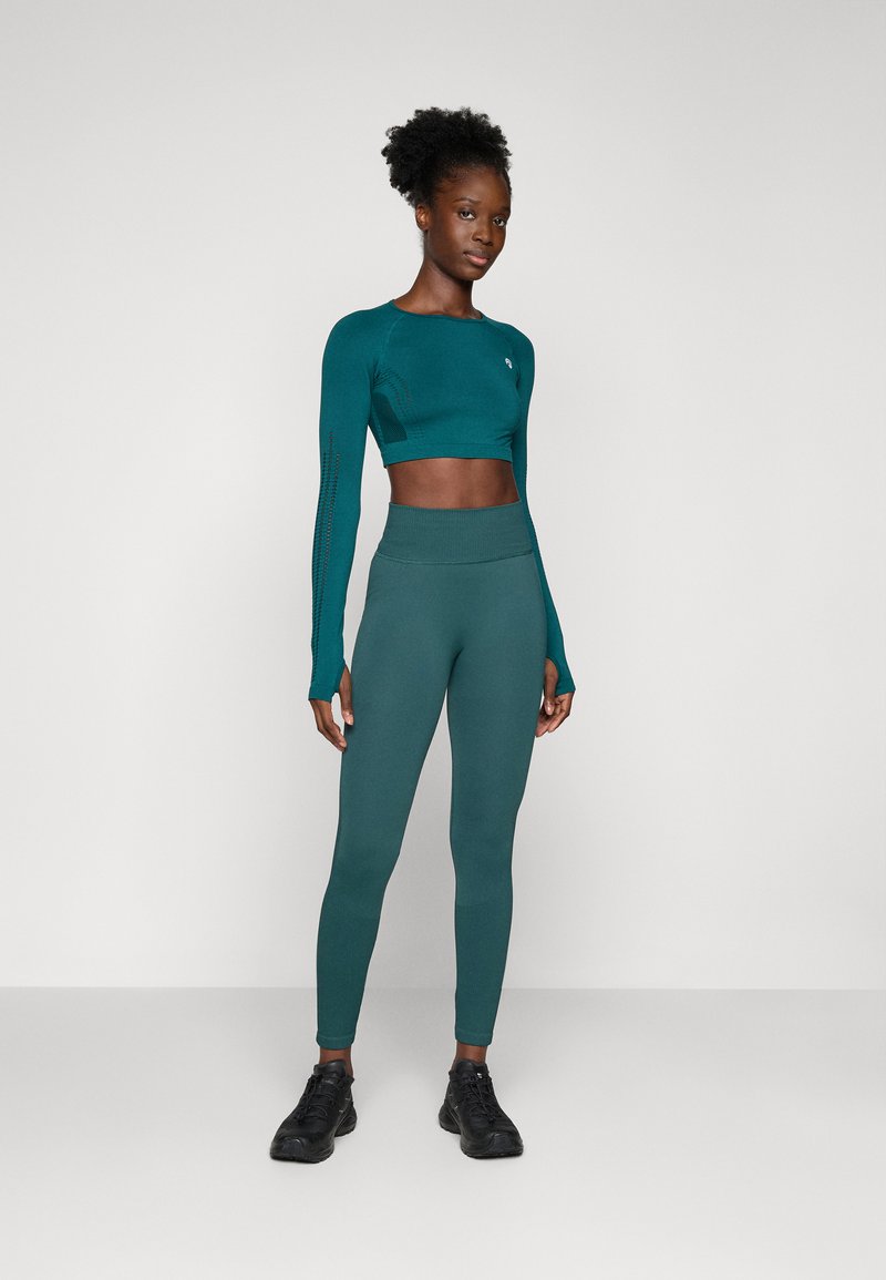 Top corto de manga larga ajustado en color azul verdoso con detalles en los paneles laterales, combinado con leggins de cintura alta en azul verdoso. Lleva puestos unos zapatos deportivos negros.
