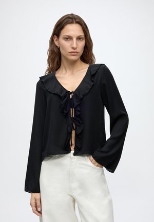 Femme portant un chemisier noir à volants avec des liens, associé à un pantalon blanc taille haute, debout avec une main dans la poche sur fond blanc.