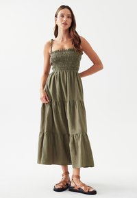 Olivenfarbiges Maxikleid mit gerafftem Oberteil, dünnen Trägern und gestuften Rüschensäumen. Kombiniert mit braunen Riemchensandalen. Schlichter Hintergrund.