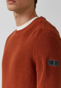 Roestkleurige gebreide trui met een ribbelstructuur, voorzien van een witte kraag en een zwart logopatch op de linkermouw.