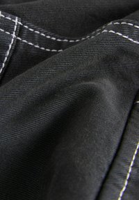 Musta kangas, jossa on hienovarainen ribbikuvio, ja jolla on kontrastina valkoiset ompeleet. Materiaali näyttää pehmeältä ja hieman valuvalta.