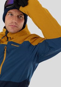 Veste colorée en jaune et bleu, fabriquée dans un matériau lisse et résistant aux intempéries. Elle dispose d'un col montant et d'une fermeture éclair ajustable.
