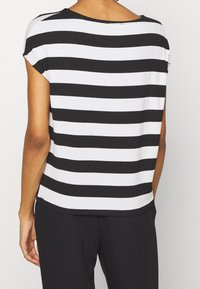 Eine Person trägt ein kurzärmeliges, schwarz-weiß gestreiftes Hemd mit horizontalen Streifen und schwarze Hosen, von hinten vor einem schlichten Hintergrund gezeigt.