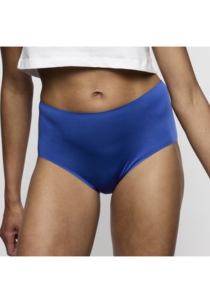 Persoon die effen blauw hoog getailleerd ondergoed en een wit cropped topje draagt, getoond van midden torso tot bovenbenen tegen een grijze achtergrond.