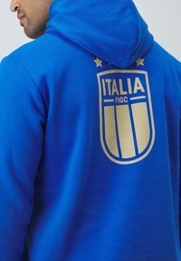 Niebieska bluza z kapturem z dużym złotym emblematem z napisem "ITALIA" i "FIGC". Projekt zawiera trzy gwiazdki nad emblematem. Wykonana z miękkiego materiału z kapturem na sznurki.