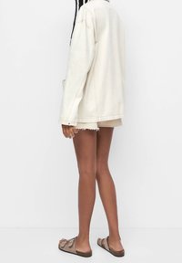 Veste en denim crème à coupe décontractée, associée à un short beige et des sandales marron. Comprend des manches retroussées et un ourlet effiloché au short.