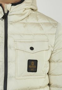 Giacca beige imbottita con cappuccio, dotata di una zip nera, una tasca sul petto con bottoni a pressione e un patch del logo in nero e oro.