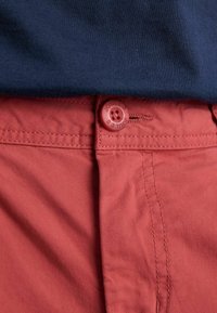 Koralo spalvos kelnės su segtukais ir matomomis siūlėmis po tamsiai mėlynu marškinėliu.