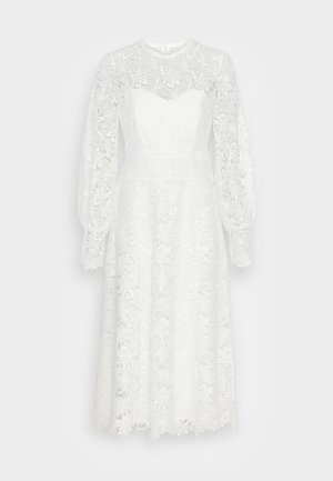 Biała sukienka do kolan z dopasowanym gorsetem, kwiatową koronkową warstwą, długimi bufiastymi rękawami i okrągłym dekoltem. Ma lekko rozszerzany dół.