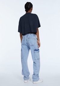 Camiseta gris oscuro cortada, combinada con pantalones cargo azul claro con múltiples bolsillos y un ajuste ligeramente holgado. Zapatillas blancas completan el look.