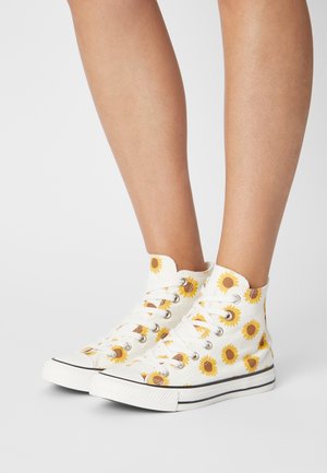 White high-top sneakers with yellow sunflower print worn on bare feet against a plain white background.