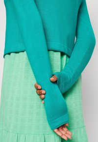Tyrkysový svetr s dlouhými rukávy, vrstvený přes mintově zelené texturované šaty. Svetr má zkrácený střih a přiléhavé manžety.