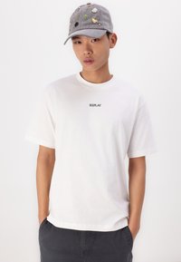 Hvid bomulds t-shirt med "REPLAY" logo på brystet, parret med en grå kasket med farverige dekorative lapper og en flad skygge.