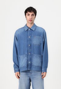 Lyseblå denimjakke med knapper i front, store frontlommer, spiss krage og lange ermer. Svakt falmet tekstur gjennom hele.