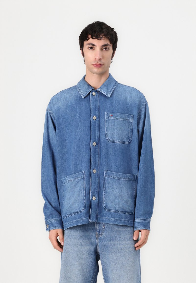 Lyseblå denimjakke med knapper i front, store frontlommer, spiss krage og lange ermer. Svakt falmet tekstur gjennom hele.