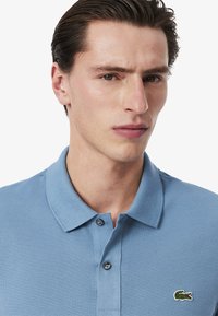 Camisa polo azul claro hecha de algodón texturizado, con un placket de dos botones y un pequeño logo bordado de cocodrilo en el pecho.