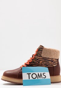 Botte d'hiver en cuir marron à rayures tigre avec des lacets orange, un col en peau lainée beige, une semelle beige et une étiquette de marque TOMS bleue.