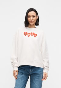 Svetlo bež jopa z okroglim izrezom, okrašena z rdečimi srcastimi poudarki in besedo "HUGO" v krepkih črkah čez prsni koš.