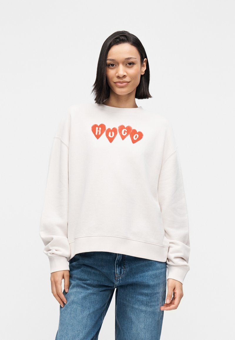 Svetlo bež jopa z okroglim izrezom, okrašena z rdečimi srcastimi poudarki in besedo "HUGO" v krepkih črkah čez prsni koš.