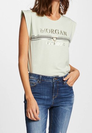 Frau trägt ein ärmelloses hellbeige Oberteil mit metallicfarbenem "MORGAN GLAMOUR" Schriftzug und Herz-Detail, kombiniert mit blauen Jeans.